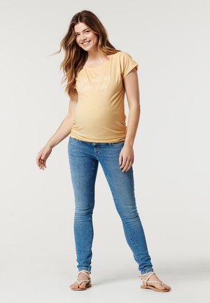 Camiseta de maternidade de manga curta amarelo claro com texto branco "Tu fazes o meu coração sorrir", combinada com jeans skinny azuis e sandálias bege.