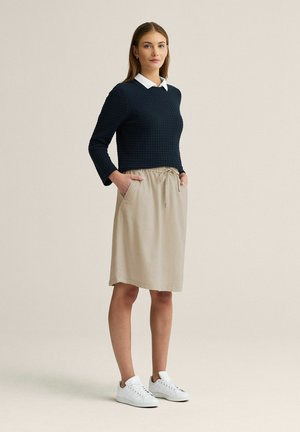 Kvinde stående med hænderne i lommerne på en beige nederdel, iført en sort struktureret sweater over en hvid skjorte med krave og hvide sneakers.
