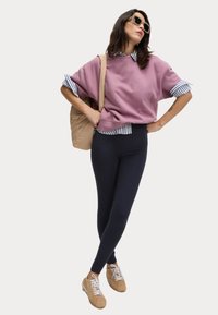 Overmaatse roze sweatshirt met een gestreept blauw-wit shirt eronder, gecombineerd met marineblauwe legging en beige sneakers. Draagt een lichtbeige rugzak.