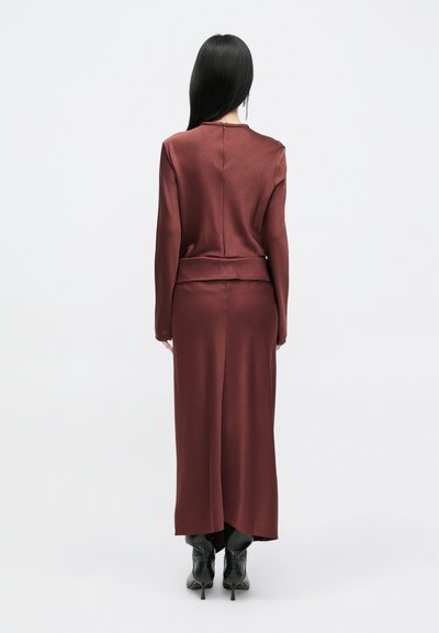 Vestido longo bordeaux com uma textura suave de cetim, apresentando mangas longas, um decote alto e uma fenda nas costas central. Combinado com botas pretas de salto.