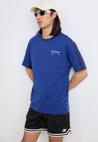 T-shirt en coton bleu avec des accents bleu marine, à manches courtes, et logo "New Balance" brodé sur la poitrine. Porté avec un short en maille noir.