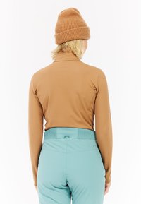 Langsleeve bruine top gecombineerd met turquoise skibroek, met daarop een gebreide tan beanie, gezien vanuit de achterkant. Glad materiaal en getailleerd ontwerp.