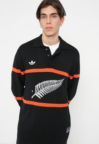 Črna pletena polo majica z naramnicami v oranžni barvi in belo grafiko praproti; vključuje zapenjanje na gumbe in adidas logo na prsih.