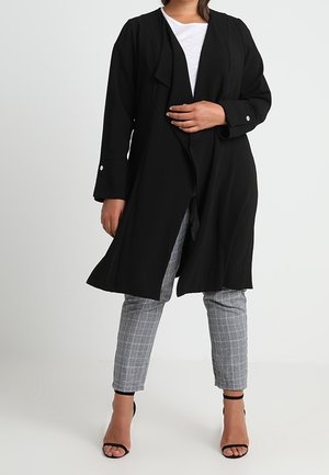 Manteau long noir au design fluide, avec des manches larges, un drapé à l'avant et des accents de boutons. Associé à un pantalon à carreaux gris et des talons.
