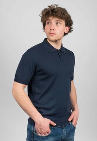 Námořnické modré pletené polo tričko s klasickým límcem, krátkými rukávy a žebrovaným lemem. Tkanina se zdá hladká s jemnou texturou.
