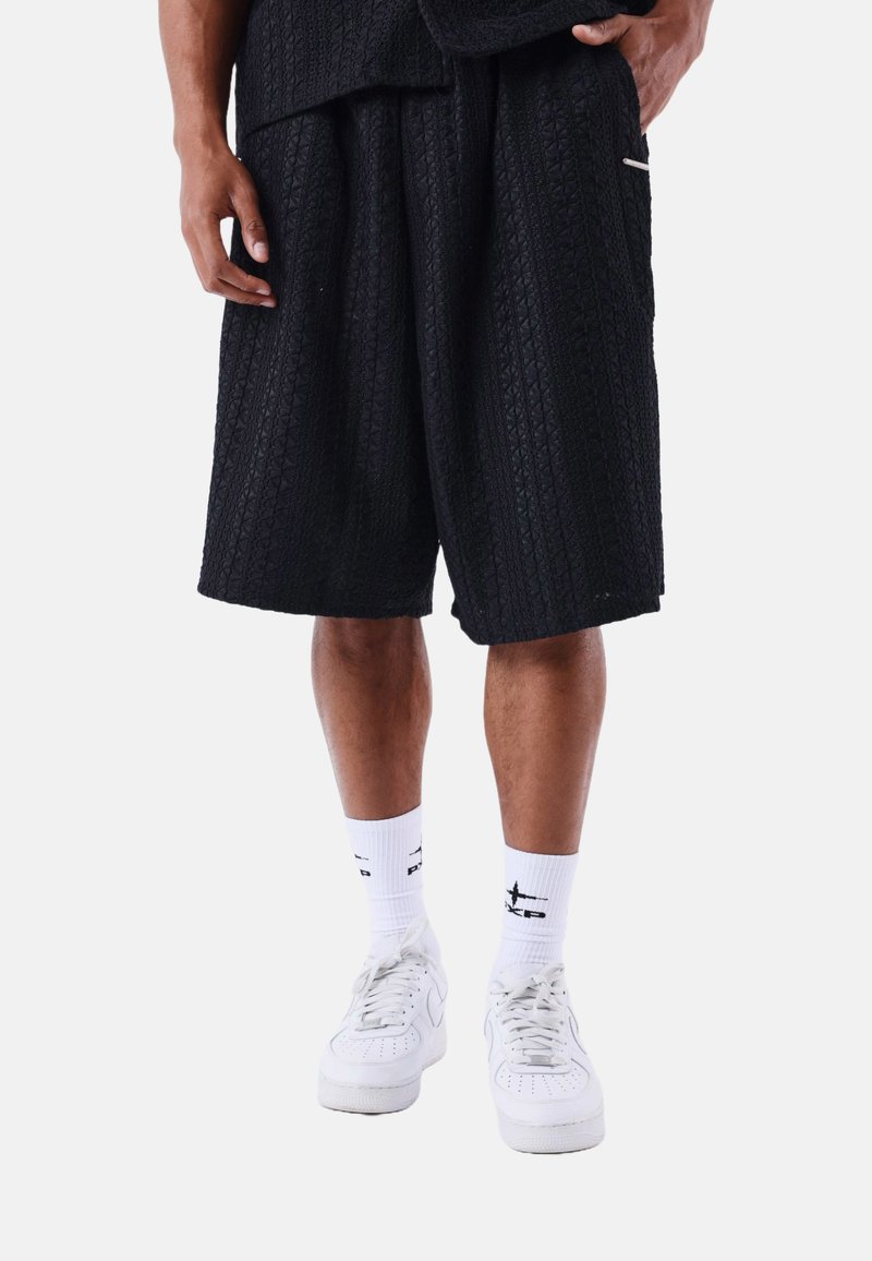 Pantalones cortos negros con textura y corte amplio, con un bolsillo lateral y un acento bordado. Combinados con calcetines blancos y zapatillas blancas.