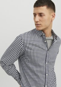 Camisa de botones a cuadros en azul marino y blanco, de mangas largas y con cuello clásico; llevada sobre una camiseta gris de cuello redondo.