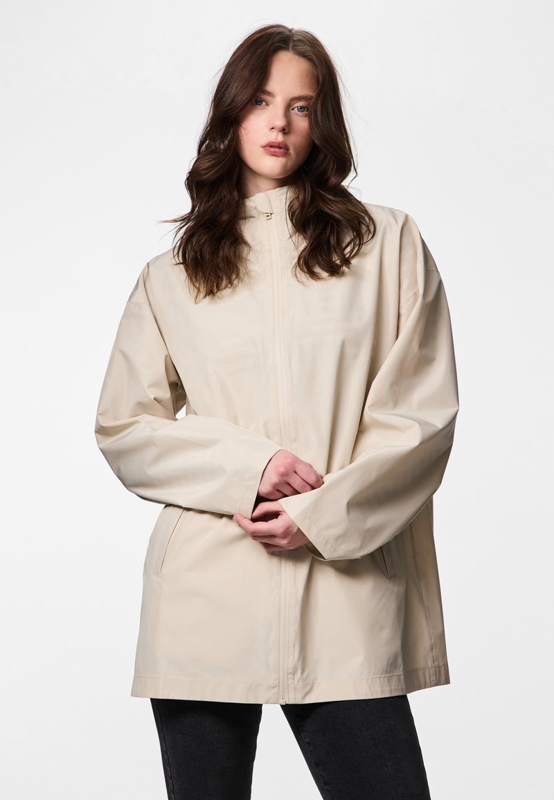 Lainelise pruuni juustega naine, kes kannab helerohelist lukuga jope ja musti pükse, seisab valgel taustal, käed rüpes ees.