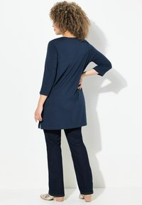 Dunkelblaue Tunika mit Dreiviertelärmeln, abgerundetem Saum und seitlichen Schlitzen, kombiniert mit dunkelblauen Jeans und beigen Freizeitschuhen.