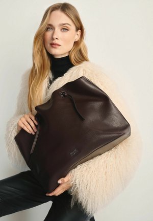 Kobieta z długimi blond włosami, w kremowej, puszystej kurtce i czarnych spodniach, trzyma dużą, ciemnobrązową skórzaną torbę na ramię.