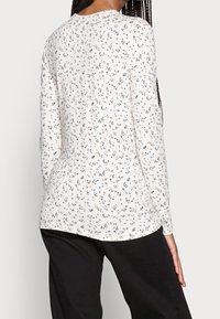 Langärmelig, crèmekleurig top met een bloemmotief in blauw, oranje en groen. Glad oppervlak en afgeronde zoom. Gecombineerd met zwarte broek.