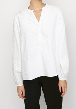 White long-sleeve blouse with a textured pattern, henley neckline, button placket, and gathered cuffs. Worn over dark pants.
