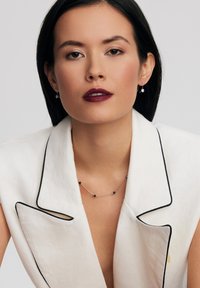 Srebrny naszyjnik z czarnymi koralikami, noszony z białym lnianym żakietem z czarnymi wykończeniami. Modelka ma proste czarne włosy i ciemną pomadkę.