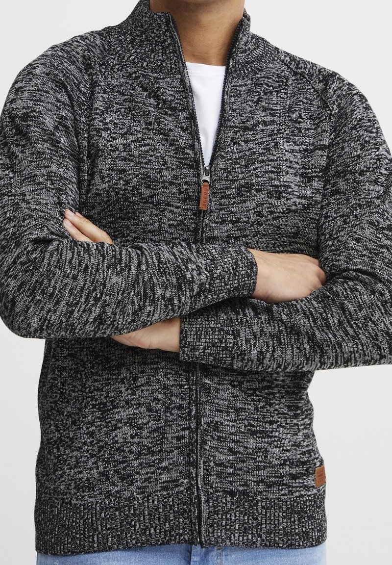 Black and gray knitted zip-up cardigan with a high collar, ribbed cuffs, and a leather tag detail at the hem.