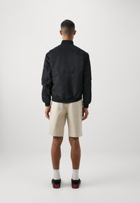 Svart bomberjakke med en glatt glans, ribbet krage og mansjetter, iført over lyse beige shorts. Svarte sko med røde detaljer på hælen.
