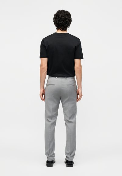 Άνδρας με σγουρά μαλλιά, γυρισμένος με την πλάτη, φοράει μαύρο κοντομάνικο μπλουζάκι, ανοιχτό γκρι παντελόνι και μαύρα παπούτσια σε απλό φόντο.