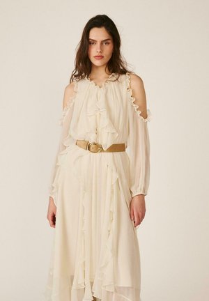 Mujer con cabello largo y oscuro, vestida con un vestido crema de hombros descubiertos con volantes, mangas largas y un cinturón beige, de pie frente a un fondo liso.