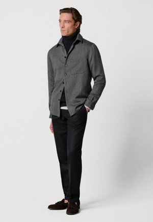 Mann trägt graues Hemd mit Knöpfen über einem schwarzen Rollkragenpullover, schwarze Hose und dunkle Loafers, steht mit einer Hand in der Tasche vor weißem Hintergrund.
