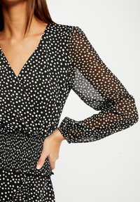 Blusa negra con lunares, escote en V, mangas largas transparentes y cinturilla fruncida, con patrones circulares blancos por todo el tejido.