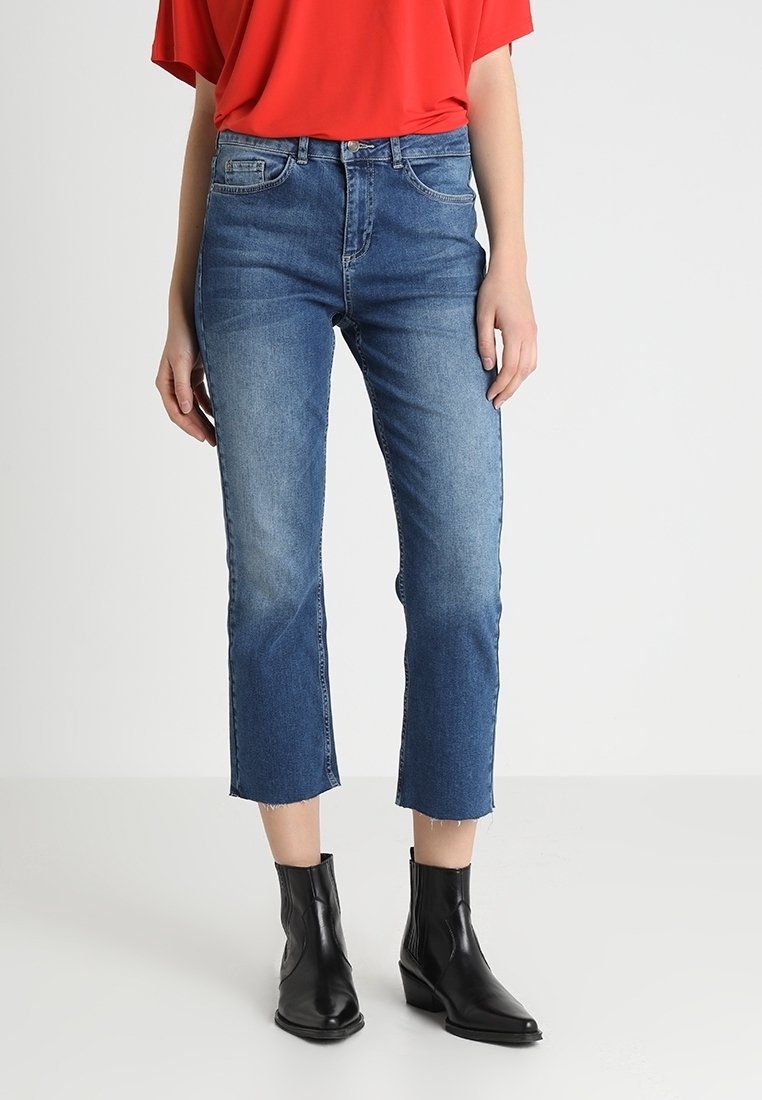 Personne portant un haut rouge, un jean bleu court et des bottines noires, debout devant un fond uni clair. Seule la partie inférieure du corps est visible.