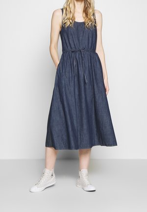 Kvinde iført ærmeløs mørk denim midi-kjole med bindebånd i taljen og hvide høje sneakers mod en ensfarvet lys baggrund.