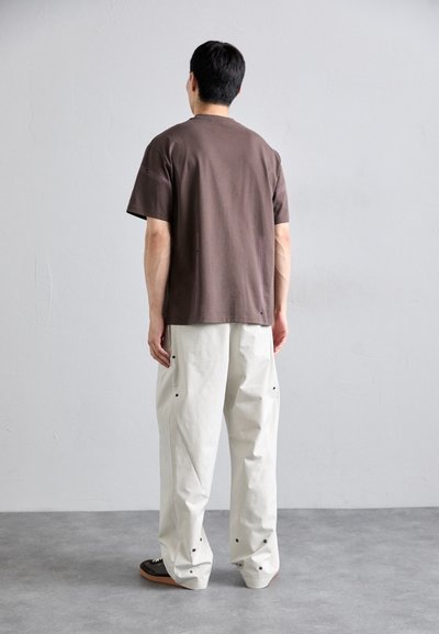 Braunfarbendes T-Shirt mit kurzen Ärmeln und lässigem Schnitt, kombiniert mit cremefarbenen, weit geschnittenen Hosen mit schwarzen Nietenakzenten und einer glatten Textur.