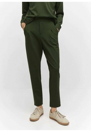 Osoba ubrana w ciemnozielone, zwężane spodnie z rękami w kieszeniach, nosząca beżowe trampki na białym tle.