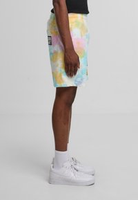 Pastelfarvede tie-dye shorts (pink, blå, gul) med en sidelomme, lavet af blødt stof, kombineret med hvide sneakers og ankelstrømper.