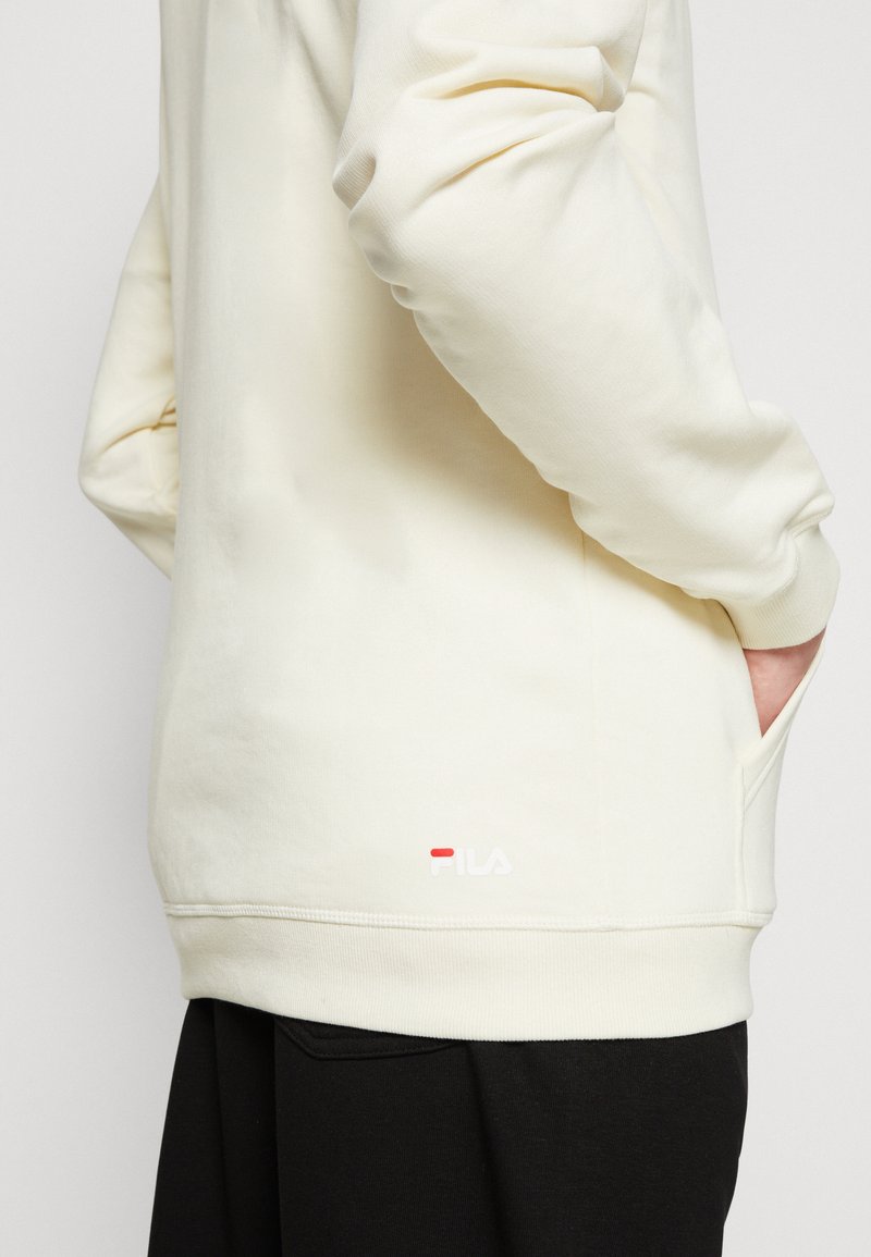 Cremefarbener Pullover aus weichem Stoff, mit lockerer Passform, seitlicher Tasche und gesticktem Logo nahe dem Saum.