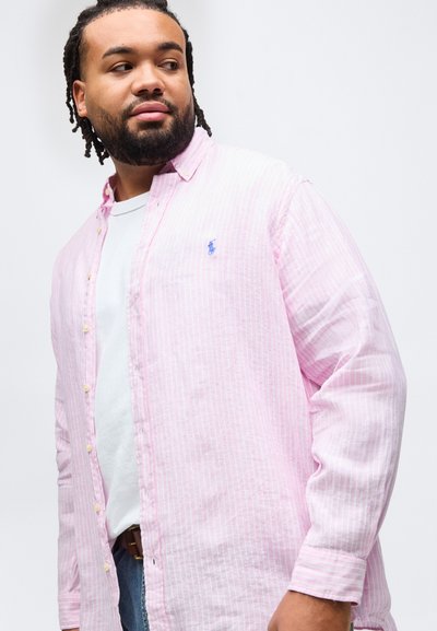 Άνδρας με γένια και dreadlocks, φορώντας ένα ανοιχτό ροζ ριγέ πουκάμισο με κουμπιά πάνω από ένα λευκό μπλουζάκι, κοιτάζει πλάγια μπροστά σε ένα απλό λευκό φόντο.