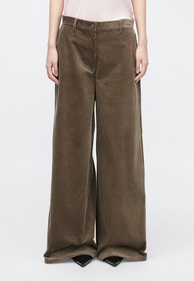 Pantalon en velours côtelé à jambes larges de couleur marron foncé, présentant une texture à côtes verticales, une fermeture à bouton sur le devant et des poches latérales, associé à des talons noirs.