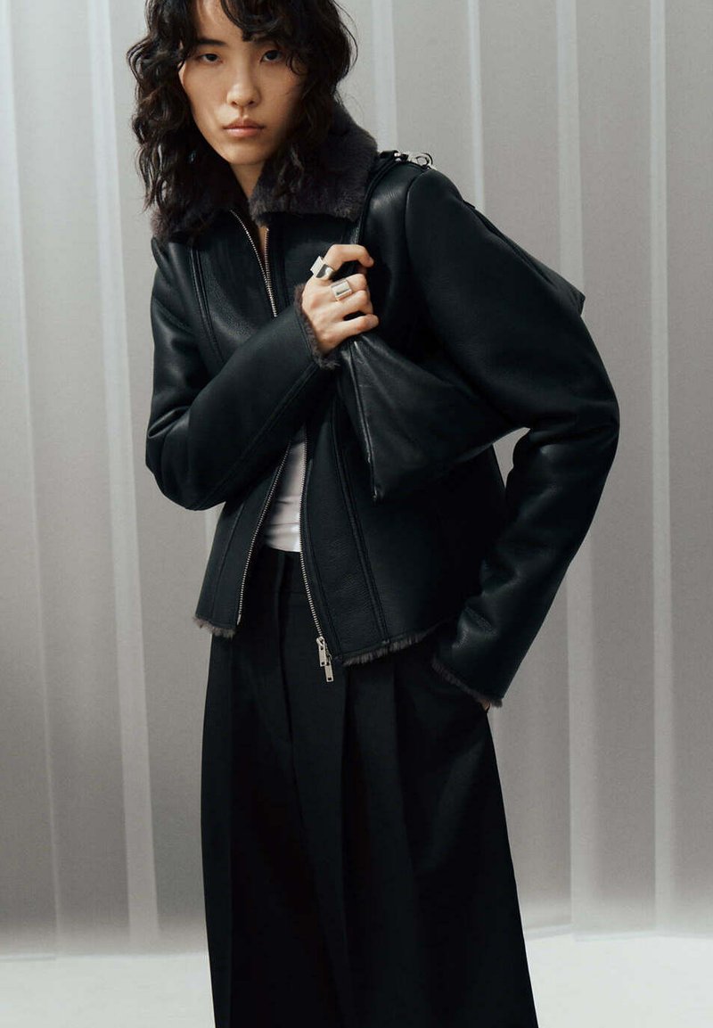 Jeune femme aux cheveux bouclés portant une veste en cuir noir en peau lainée, un pantalon large noir et tenant un sac à main noir triangulaire.
