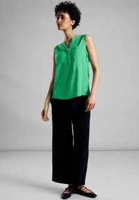 Vihreä hihaton toppi, jossa on napitettava V-pääntie ja kaksi rintataskua, yhdistettynä löysiin mustiin housuihin ja tasapohjaisiin kenkiin. Sujuva rakenne.