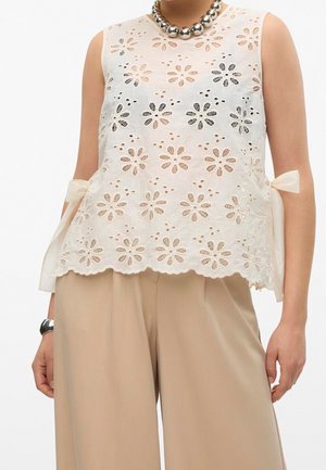 Biała, bezrękawnikowa bluzka z koronką, ozdobiona kwiatowymi wycięciami i kokardami po bokach, zestawiona z beżowymi spodniami o szerokich nogawkach oraz masywnym srebrnym naszyjnikiem.