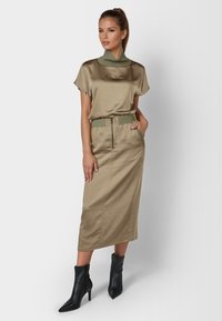 Satinkleid in Khaki mit kurzen Ärmeln und hohem Kragen, kombiniert mit einem knielangen Satinenrock mit Reißverschluss und grünem Bandelement. Schwarze spitze Stiefel.
