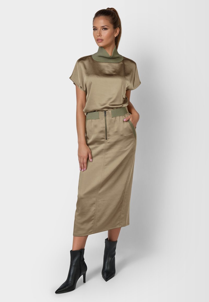 Satinkleid in Khaki mit kurzen Ärmeln und hohem Kragen, kombiniert mit einem knielangen Satinenrock mit Reißverschluss und grünem Bandelement. Schwarze spitze Stiefel.