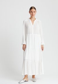 Robe longue blanche à niveaux avec un col, en tissu texturé, à manches longues, portée avec des chaussures plates argentées à bride arrière.