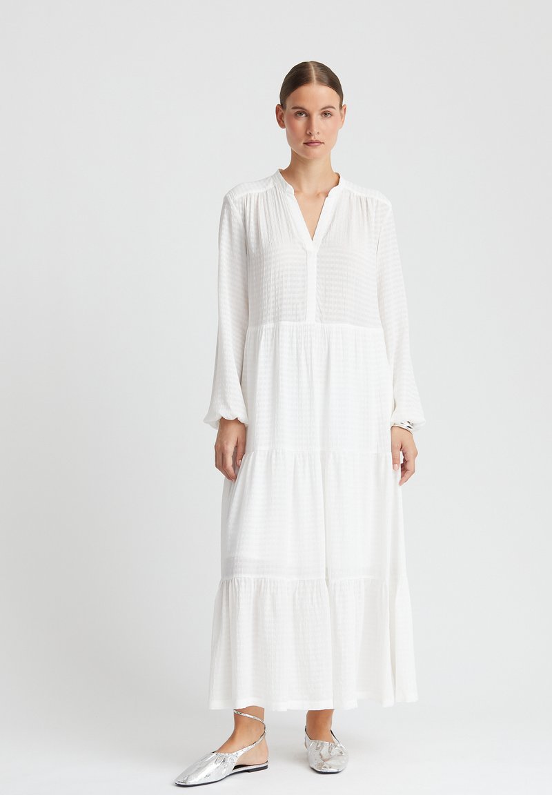 Robe longue blanche à niveaux avec un col, en tissu texturé, à manches longues, portée avec des chaussures plates argentées à bride arrière.