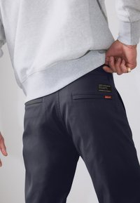 Pantaloni bleumarin închis cu o textură netedă, având un buzunar pe spate cu o etichetă de marcă. Purtați cu un hanorac gri deschis.