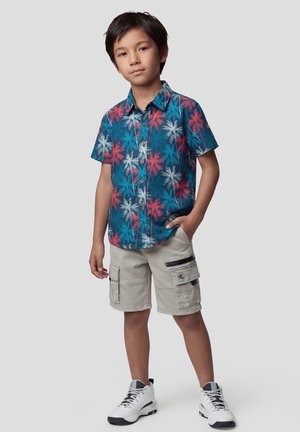 Niño de pie con una mano en el bolsillo, vistiendo una camisa azul con estampado tropical, pantalones cortos cargo beige y zapatillas blancas contra un fondo liso.