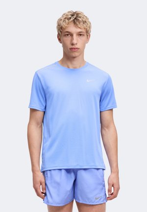Giovane uomo con capelli corti e ricci biondi che indossa una t-shirt Nike azzurro chiaro e pantaloncini abbinati, in piedi davanti a uno sfondo chiaro uniforme.