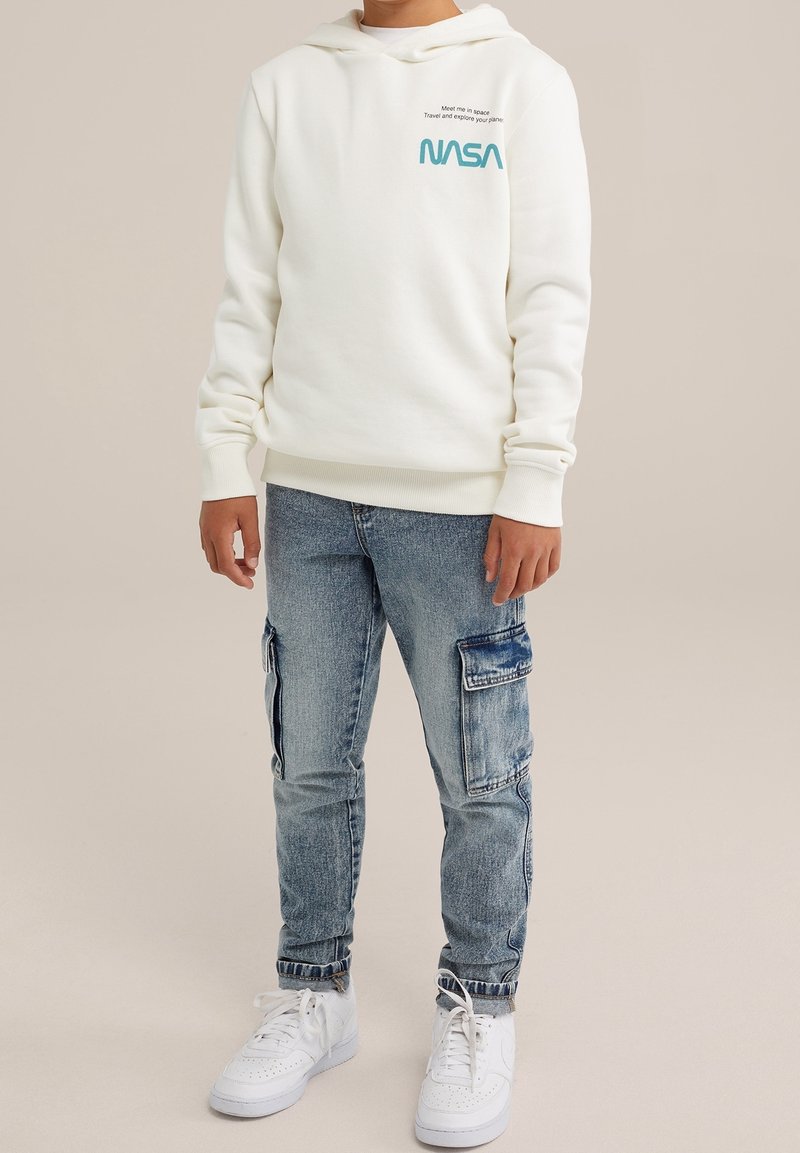 Sweat à capuche blanc avec le logo NASA, doté d'une poche avant et de poignets côtelés. Associé à un jean cargo bleu clair déchiré et des baskets blanches.