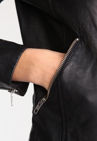 Giacca in pelle nera con tasca con zip e dettagli in metallo argentato. Vista ravvicinata che mette in evidenza la texture liscia e il design aderente.