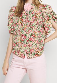 Blouse à imprimé floral en rose doux avec des manches bouffantes, un devant boutonné et un accent de rose décoratif au niveau du col. Associée à un pantalon rose pâle.