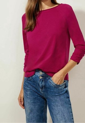 Femme portant un top à manches longues magenta avec des manches froncées et un jean en denim bleu, une main dans la poche, debout devant un fond uni.