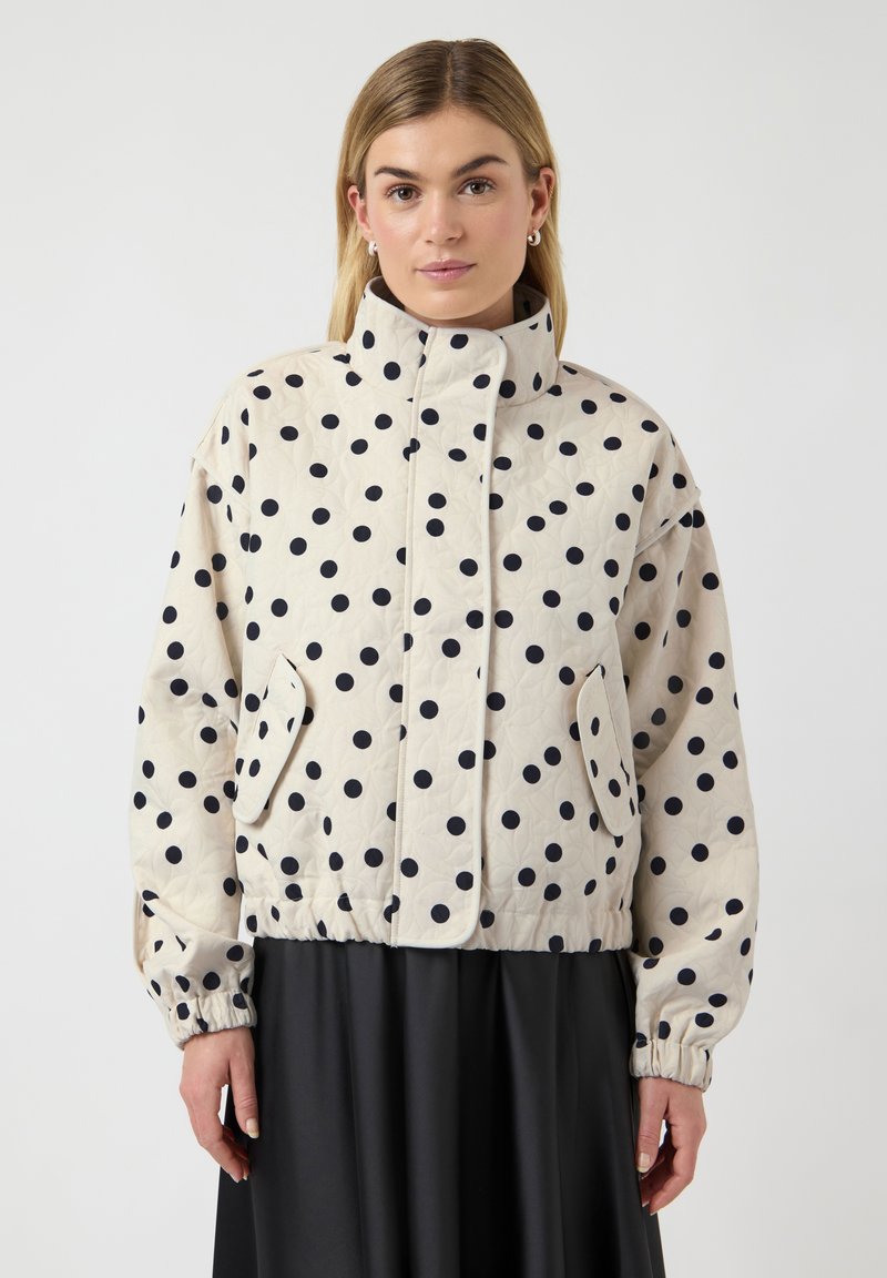 Femme portant une veste matelassée beige à pois noirs, col montant, poches à rabat et poignets élastiques, associée à une jupe noire.
