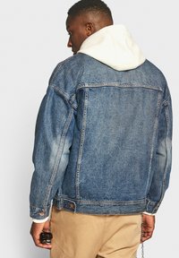 Donkerblauwe denimjack met een relaxte pasvorm, voorzien van een knoopsluiting en twee borstzakken. Gelaagd met een crème kaptrui eronder.