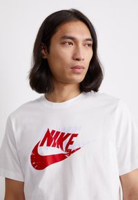 Balta medvilnė marškinėlių su dideliu raudonu ir baltu nuleistų spalvų Nike logotipu per krūtinę. Apskritas iškirpimas ir trumpomis rankovėmis.