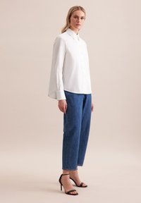 Biała koszula z wysokim kołnierzykiem, zestawiona z luźnymi niebieskimi dżinsami i czarnymi sandałami na wysokim obcasie. Gładki materiał, swobodny krój.
