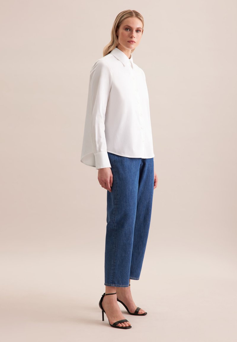 Biała koszula z wysokim kołnierzykiem, zestawiona z luźnymi niebieskimi dżinsami i czarnymi sandałami na wysokim obcasie. Gładki materiał, swobodny krój.